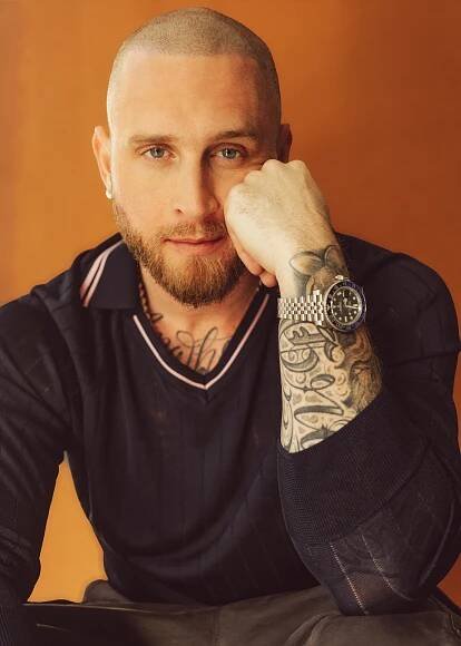 A man with a shaved head and a beard rests his chin on his hand, wearing a dark sweater. He has visi...