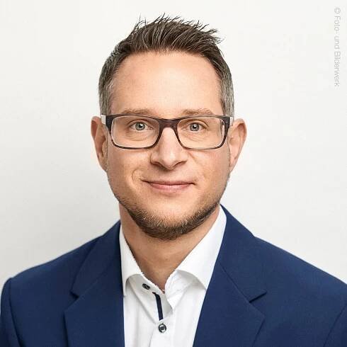 Mann lächelt freundlich direkt in die Kamera vor neutralem Hintergrund
