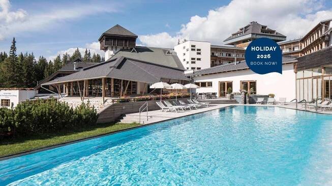 Schwimmbad vor einem Hotel, umgeben von Bäumen und Liegen, mit Text HOLIDAY 2026 BOOK NOW!