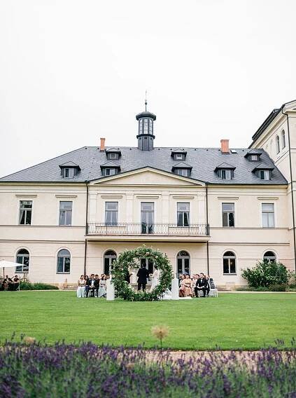 Chateau Mcely facade with the ceremony and floral arch in the foreground, lavender fields and elegant guests completing the view.