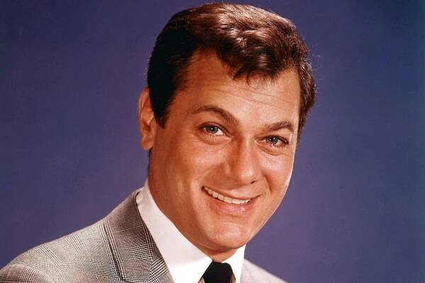 Tony Curtis (1925-2010), US actor, smiling, wearing a grey jacket, white shirt and black tie in a studio portrait, against a blue background, circa 1960. 