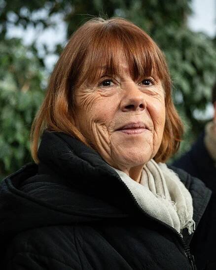 Ms. Pelicot is seen in close-up, smiling. She wears her light brown hair in a short bob. She has on a black zip-front jacket with a white scarf at her neck. 
