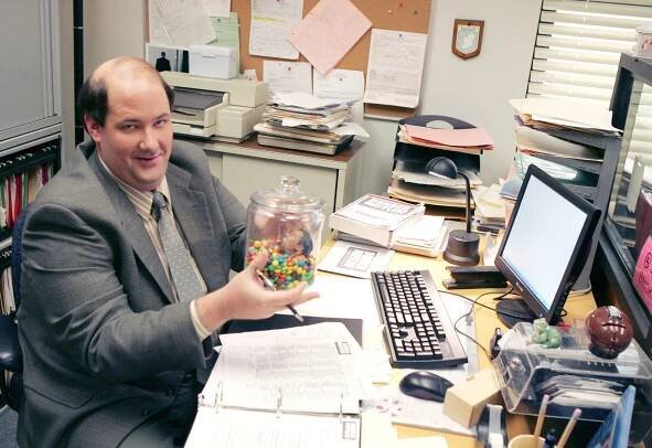 Kevin Malone from *The Office* sits at his desk, smiling and holding a jar of M&Ms.