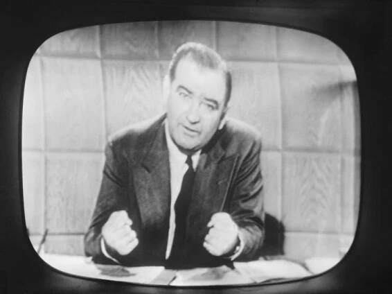 A white man sits behind a desk and is speaking in front of a telivision camera.