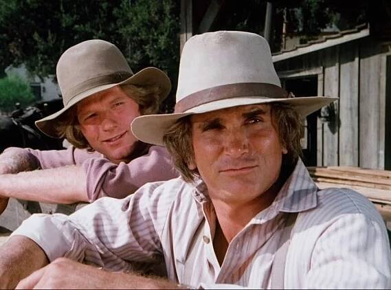Michael Landon and Dean Butler, dressed in vintage Western attire, lean on a wooden fence in an outdoor setting