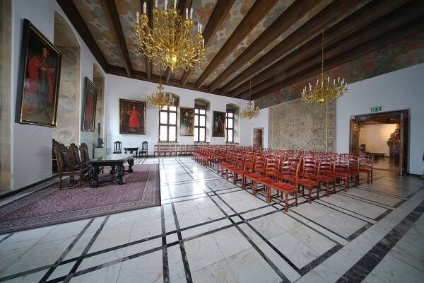 Wielka Sala Wety w Ratuszu Głównego Miasta. Duże pomieszczenie, w którym odbywają się uroczystości. Po prawej krzesła ustawione w rzędach dla widowni. Na ścianach wiszą obrazy polskich królów.