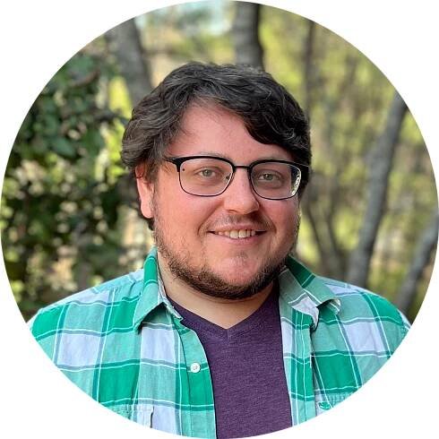 A headshot of an adult man with a beard and glasses. He is wearing a green and white checkered shirt over a purple shirt.