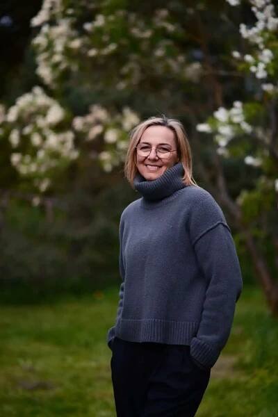 Usměvavá žena (Lenka Čápová) v šedém svetru stojící na zahradě před rozkvetlými keři.