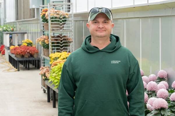 A man in a green sweatshirt smiles near flowers in a greenhouse.