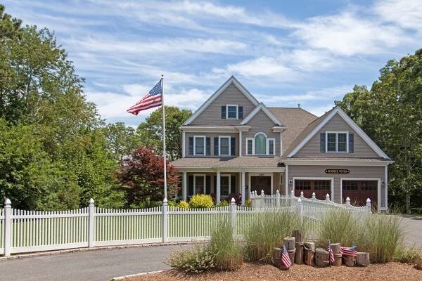 A large two-story house with beige siding and a three-car garage sits behind a white picket fence. An American flag is on a pole in the front yard. Trees and bushes surround the property under a partly cloudy sky.