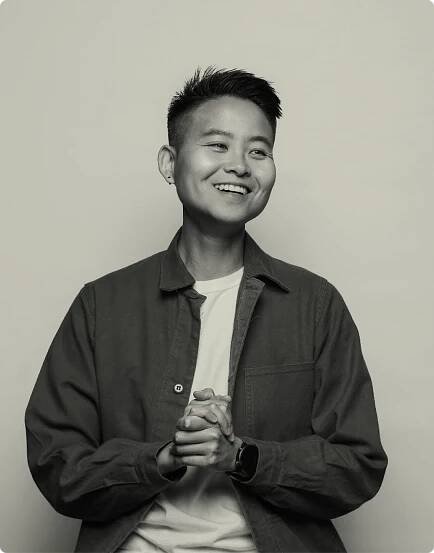 A black and white professional headshot photograph of Katie Chen. She is smiling warmly at the camera. She is wearing an unbuttoned dark overshirt with a white t-shirt underneath, a small earring, and a smartwatch. Her hands are clasped together in front of her chest. Her hair is styled in a modern short cut. The image is shot against a plain light background, creating a clean, professional look.