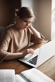Free photo focused good-looking caucasian female entrepreneur in trendy glasses, leaning head on hand and gazing at laptop screen, making notes in notebook and book, working hard.
