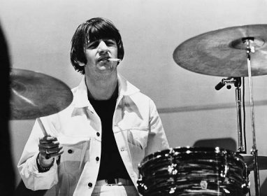 The Beatles' Ringo Starr holds a cigarette in his mouth and plays drums.