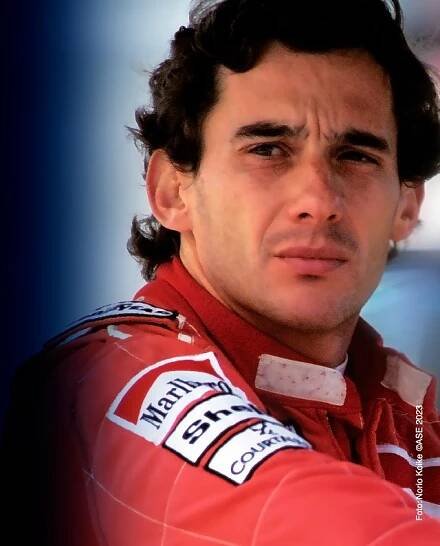 Close-up portrait of a male race car driver in a red racing suit with sponsor logos, looking contemplative amidst home decor.