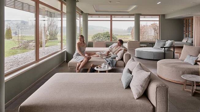 Zwei Personen entspannen auf bequemen Sofas in einem hellen, modern eingerichteten Raum mit großen Fenstern und Ausblick.