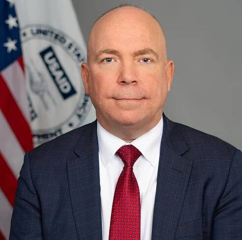 An official government portrait of Jason Gray wearing a suit in front of an American flag and USAID flag.