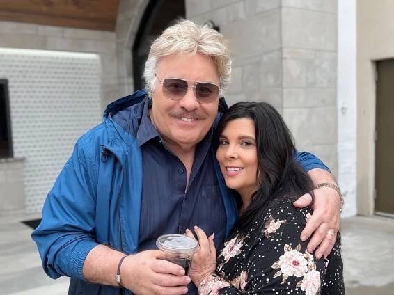 Tony Orlando, a well-known musician, is smiling and posing for a close photo with a woman. They are standing outdoors in a casual setting, with a modern stone building in the background.