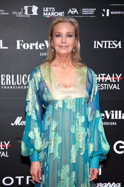 A person in a flowing, patterned gown is on a red carpet at a film festival event, standing in front of a backdrop with numerous sponsor logos