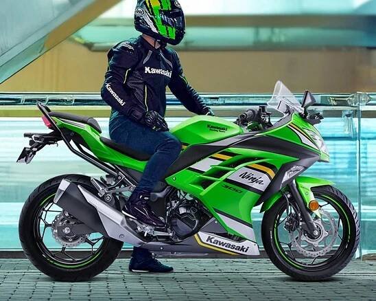 A person wearing a black and green Kawasaki motorcycle jacket, matching helmet, gloves, and dark pants is seated on a bright green Kawasaki Ninja 300 motorcycle. The motorcycle features black and white accents with yellow stripes, and has a sporty design with visible disc brakes and a large exhaust pipe. The background is an indoor setting with glass and metal railings.
