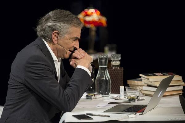 Bernard-Henri Lévy donne une représentation de sa pièce, il est seul assis derrière un bureau sur lequel il y a des livres, des boissons et un ordinateur.