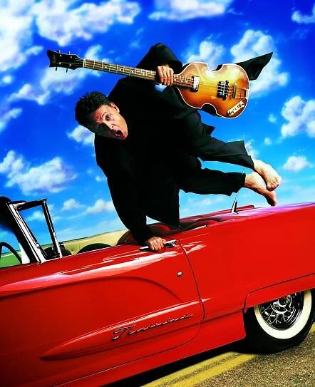 Paul jumping over a red car, holding a bass guitar