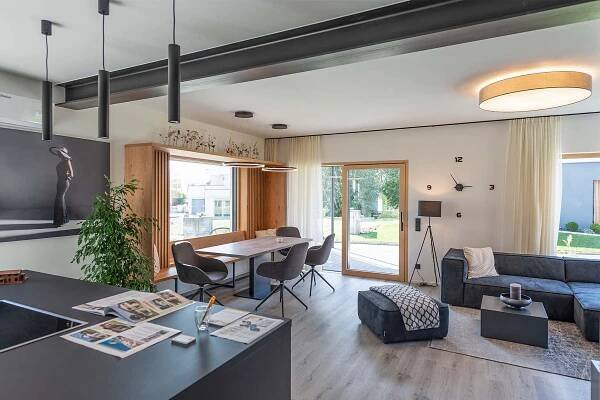 Innenansicht eines modernen Fertighauses Viden 141 von WOLF Haus. Das Wohnzimmer zeigt ein elegantes offenes Design mit einer modernen Küche, einem Esstisch mit Stühlen und einem bequemen Sofa. Große Fenster bieten reichlich Tageslicht, und eine stilvolle Wanddekoration ergänzt die ästhetische Raumgestaltung.