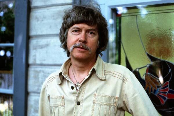 A man with shaggy dark hair and a graying mustache poses for a portrait. 