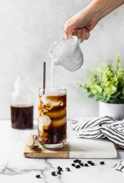 Pour shot of pouring cream into a glass of cold brew sitting on a white marble and brown wood cutting board. 