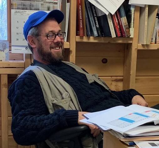A smiling white man with dark hair, a beard, glasses and wearing a blue baseball cap, a khaki vest over a bulky sweater leans back in a chair in front of a desk covered with books and papers.