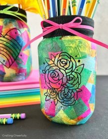 A mason jar covered in colorful pieces of tissue paper; a black vinyl rose design decorates the front of the jar. 