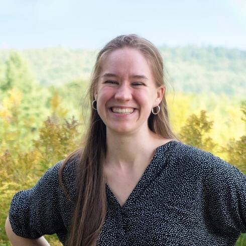 Sarah, seen from the torso up, smiles into the camera in front of a sunlit field and hills.