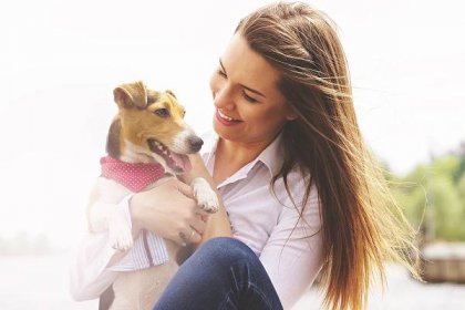 Me and my lovely pet. Portrait of young attractive woman in casual wear looking at dog and smiling while keeping dog on her hands sitting on pier.
