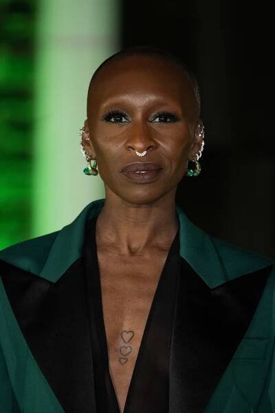 Cynthia Erivo with short hair and multiple earrings wears a stylish dark blazer with a plunging neckline, displaying heart tattoos on their chest