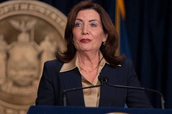A close-up of Gov. Kathy Hochul as she stands in front of a dark blue background and a giant representation of the state seal of New York.