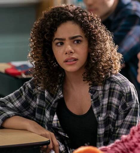 Person with curly hair in a plaid shirt looks concerned while sitting in a classroom setting