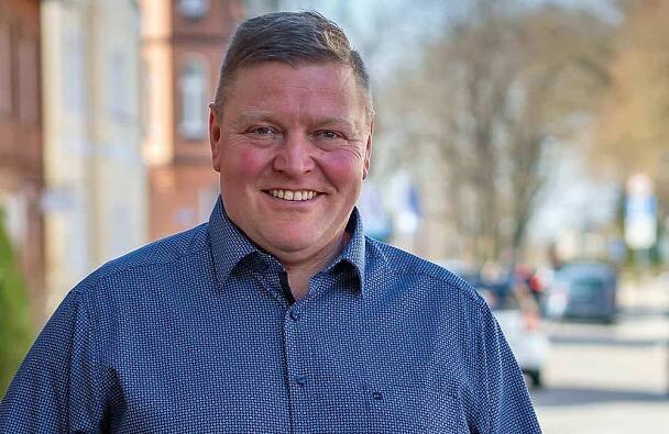 Ein Mann in einem blauen, gemusterten Button-up-Hemd steht im Freien und lächelt in die Kamera. Der Hintergrund ist eine sonnige Straße mit unscharfen Gebäuden, Bäumen und Autos.