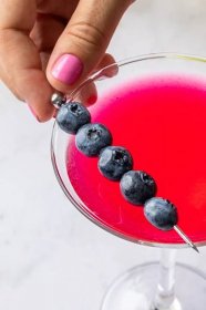 A close up of a blueberry lemon drop martini being garnished with a skewer of blueberries. 
