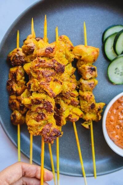 Chicken satay skewers on a blue plate with cucumber slices and peanut sauce.