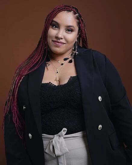 Professional portrait of performer with burgundy braids wearing black blazer against brown backdrop.
