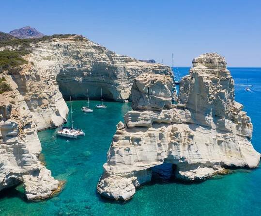 A picturesque view of boats floating on a body of water, with the island of Milos visible in the background. The scene showcases a natural landscape featuring cliffs and coastal formations under a bright sky.