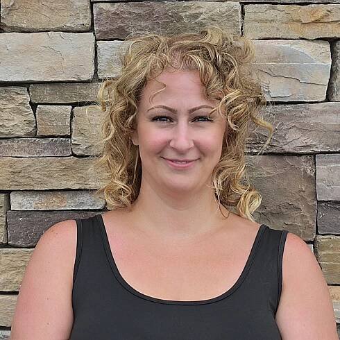 A woman with curly hair is smiling in front of a stone wall.