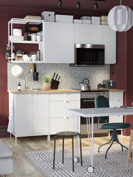A white ENHET kitchen combination along a wall in a studio apartment with a desk in front and a bed behind the wall.