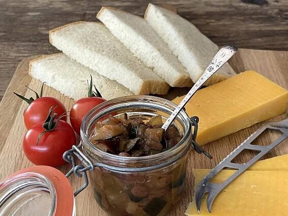 A jar of chutney with a spoon, a knife, tomatoes, cheese and bread.