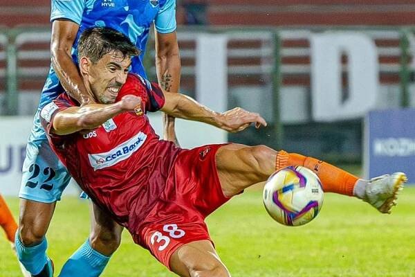 A man in a red shirt swivels to kick a soccer ball with a man in a blue shirt holding his shoulders behind him.