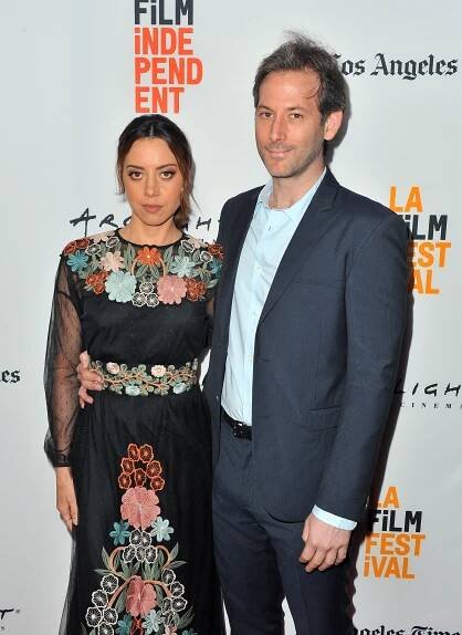 Two individuals pose on the red carpet at a film festival. The woman wears an embroidered floral dress, and the man is in a suit