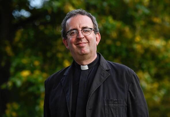 a man wearing glasses and a clergy collar smiles for the camera