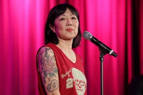 Margaret Cho with short hair, wearing a sleeveless top, speaks into a microphone, with floral tattoos on her arm