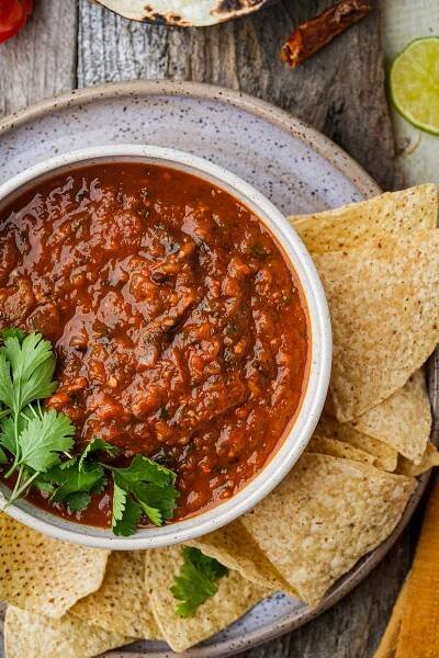 A bowl of red salsa roja garnished with cilantro leaves, surrounded by tortilla chips, placed on a rustic wooden surface.