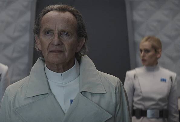 A man in a futuristic blazer in the foreground and a woman in a white futuristic belted uniform in the background; still from "Andor."