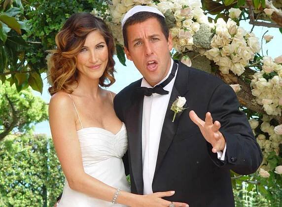 Jackie Sandler in a spaghetti strap wedding dress with her hair curled and Adam Sandler in a tuxedo, standing in front of a floral archon their wedding day 